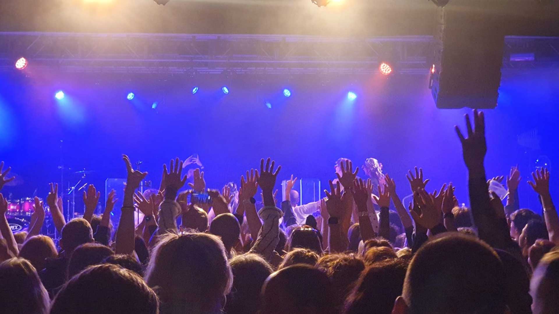 A band playing on a stage with an audience standing in front with their arms held up in the air.