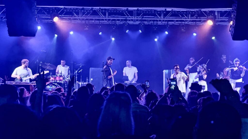 Band on a blue-lit stage with a lighting truss above and the crowd in front
