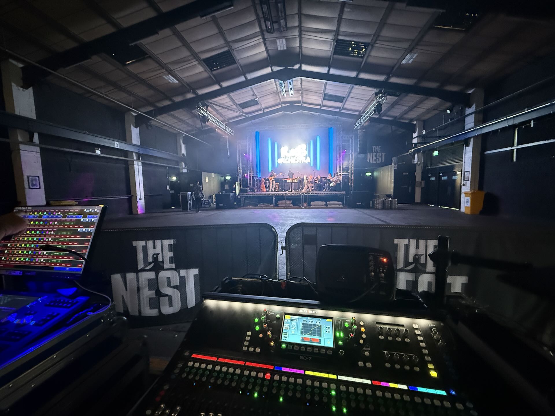 A sound desk in a large warehouse with a stage at the end. A band are playing on the stage.