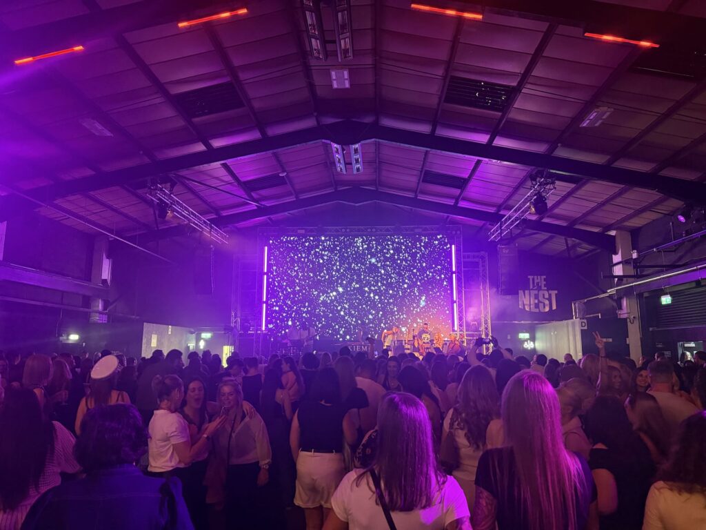 Packed crowd in a large hall lit in purple and pink