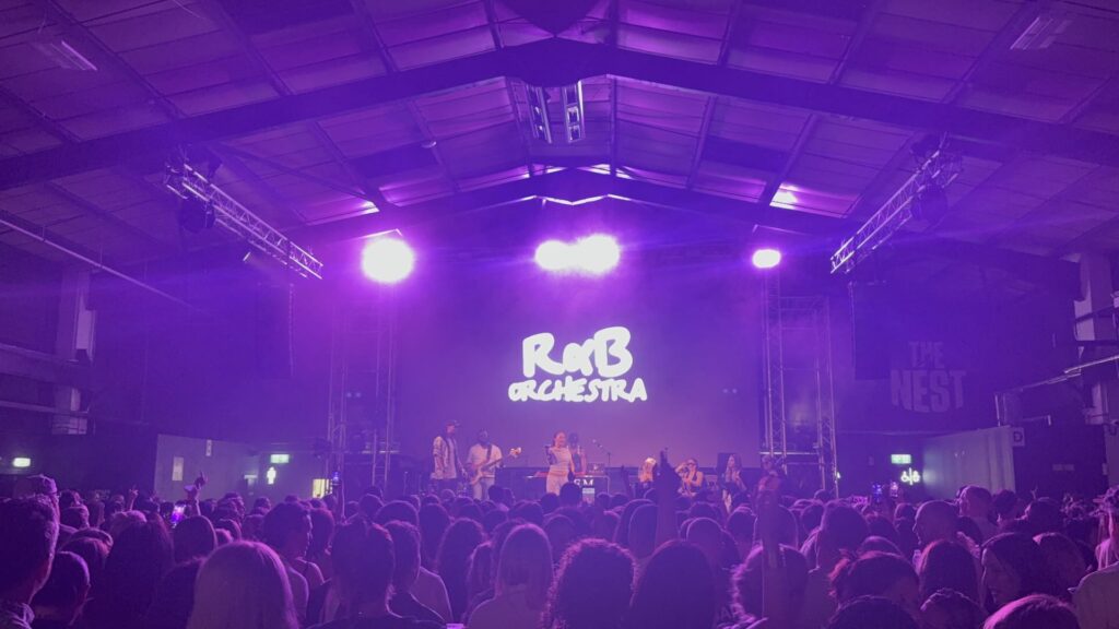 Stage lit in purple with the orchestra logo on screen and the crowd in front