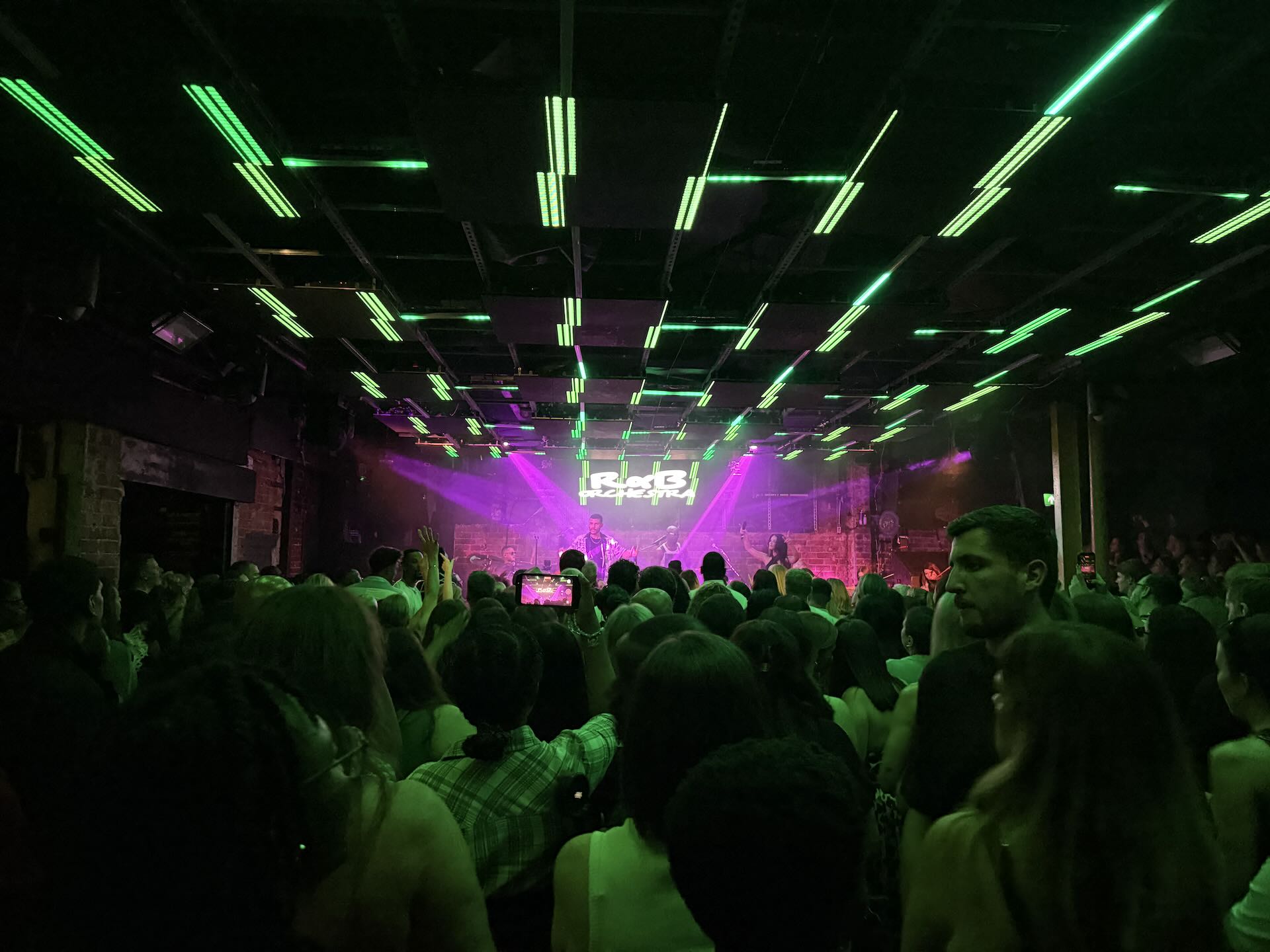 An audience of pwople stand and watch a band playing on a stage. There are green lights shinning from the stage onto the audience.