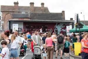 A street full of people.Some are watching a band play on a stage at the top of the street.