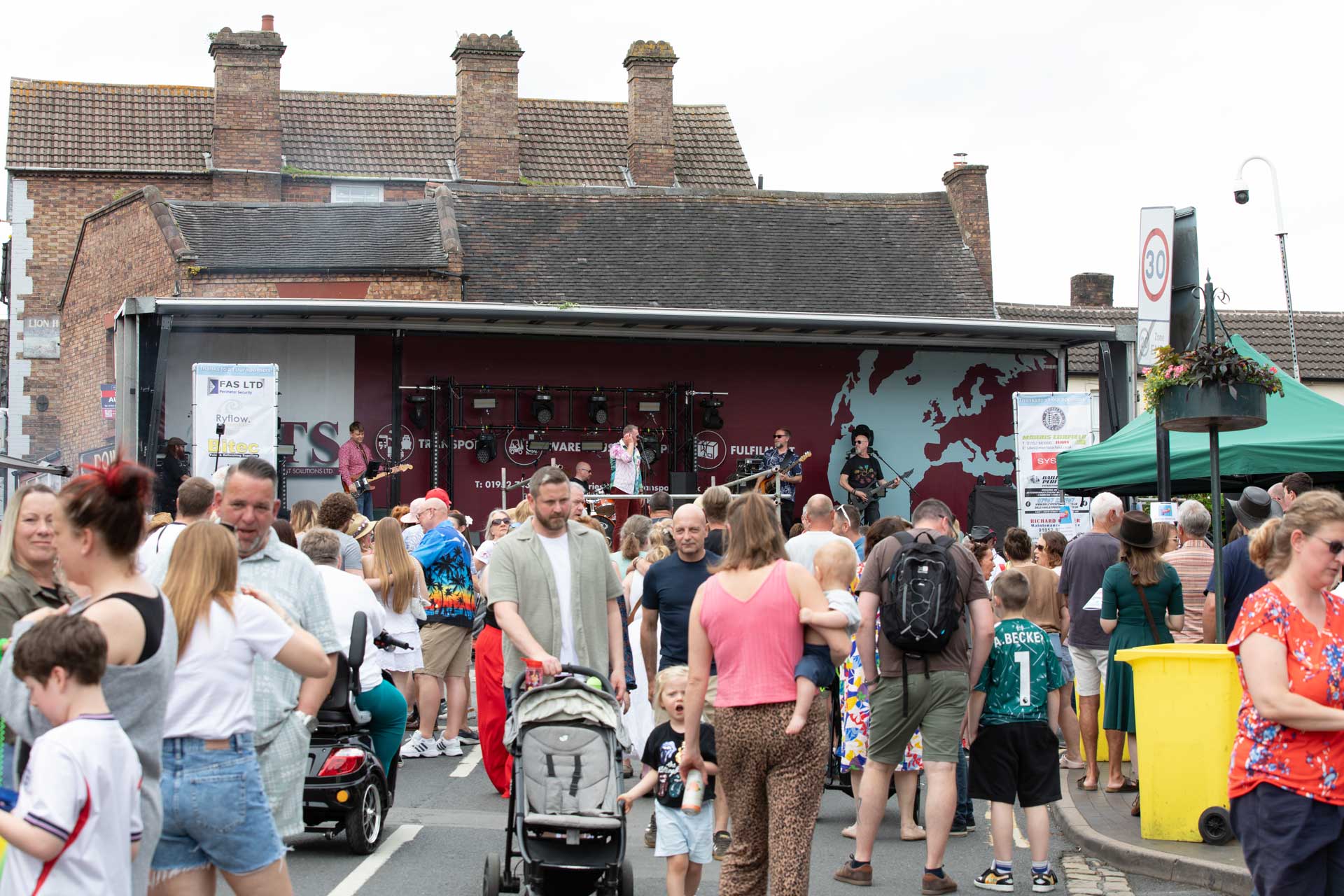 A street full of people.Some are watching a band play on a stage at the top of the street.