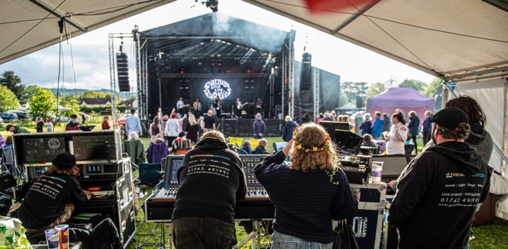 Crew and audience on the grass in front of a covered stage at Meadowfest