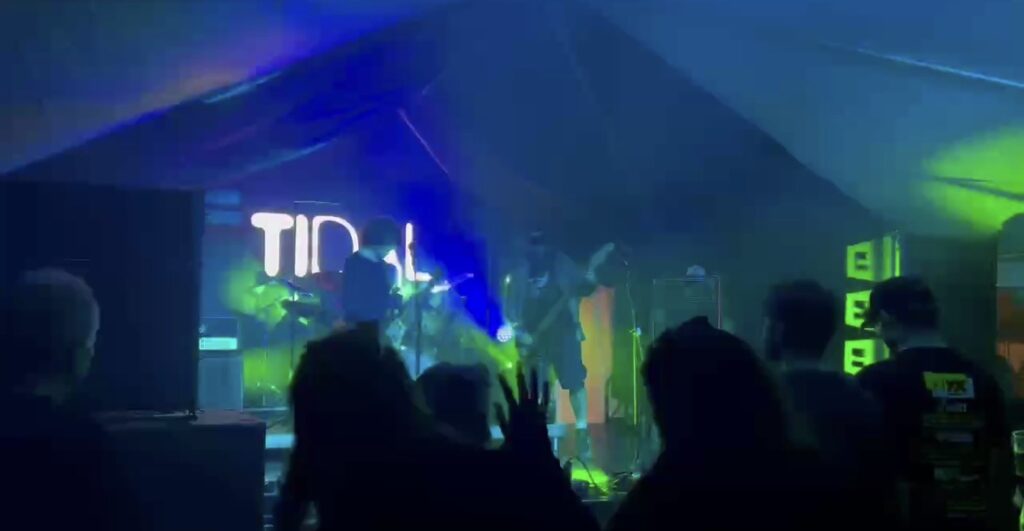 Stage lit in blue and green with a logo on the screen and the crowd in front