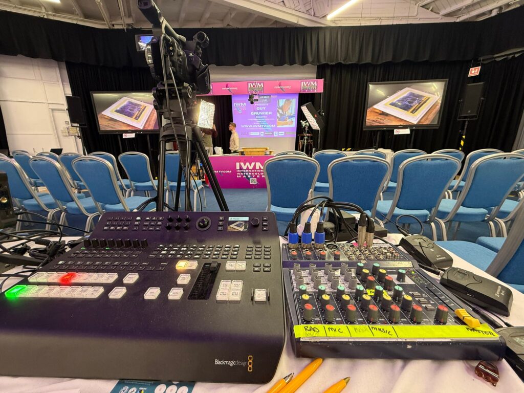Video mixing desk in the foreground with rows of chairs and screens in the hall beyond