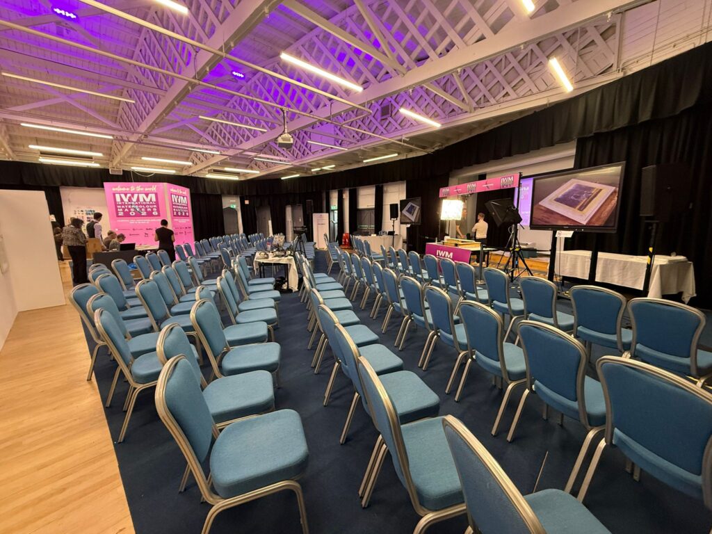 Rows of chairs set out in a hall with a lit stage and ceiling lights