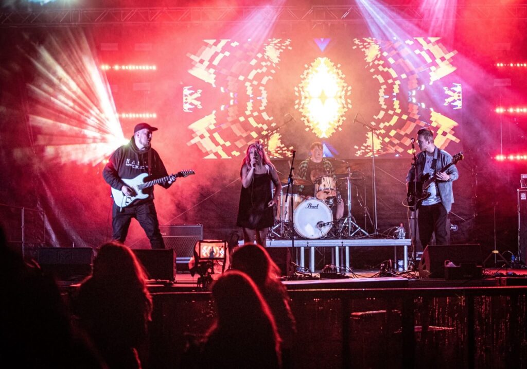 Band playing on a stage with red and orange lighting and a glowing backdrop at Meadowfest