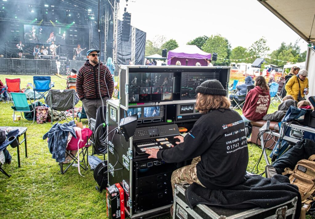 Technician at a sound desk on the grass with the festival crowd behind at Meadowfest