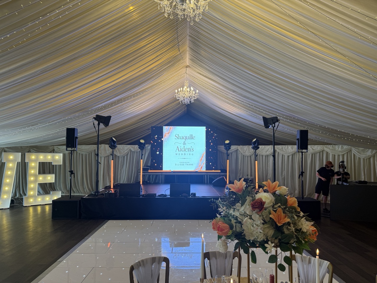A room with white material draped accross the ceiling with a stage at the end.There is a video wall on the stage that says Shaquille and Aiden's Wedding.