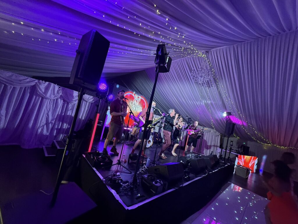 Band on a stage in a marquee with white drapes lit in purple, speakers flown at either side