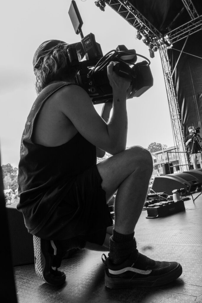 Black and white photo of a camera operator crouching with a broadcast camera