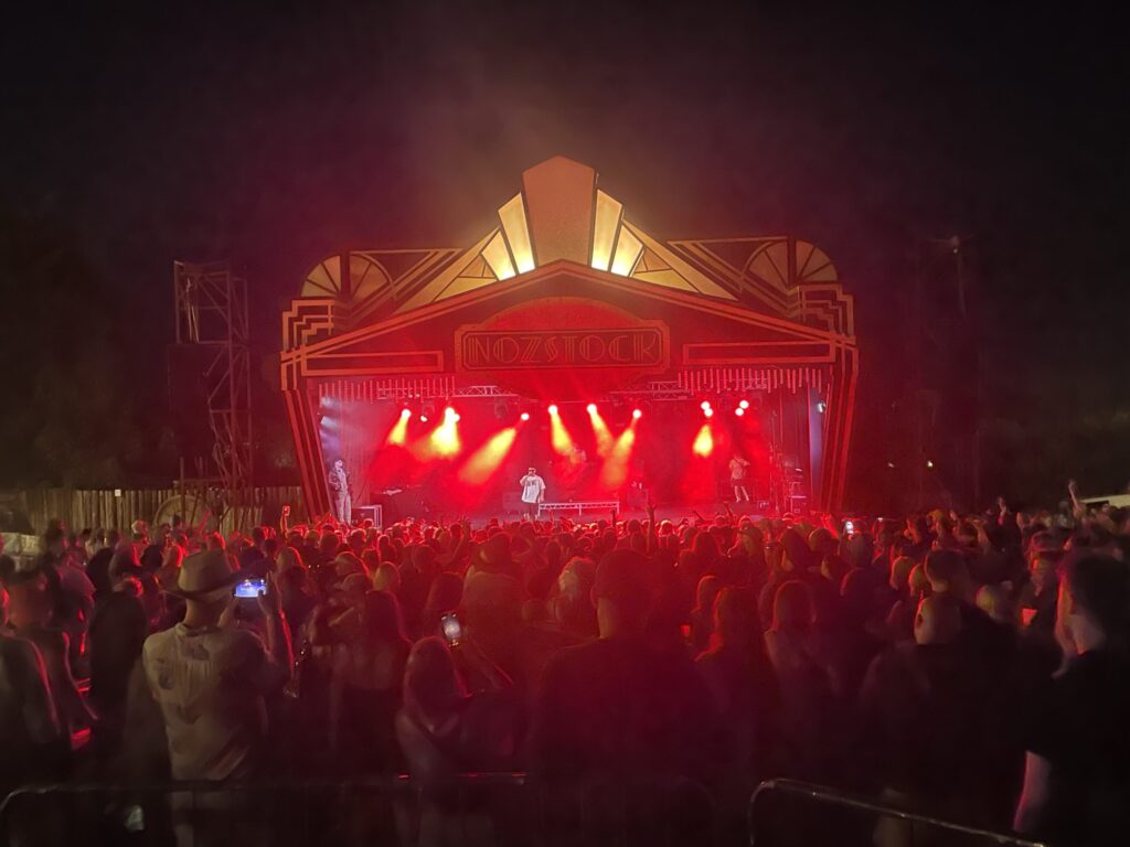 Festival stage with a canopy lit in orange and red and a crowd in front at night