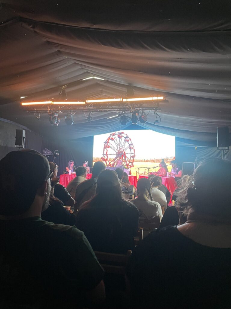 Audience facing a stage with a glowing screen under a marquee roof