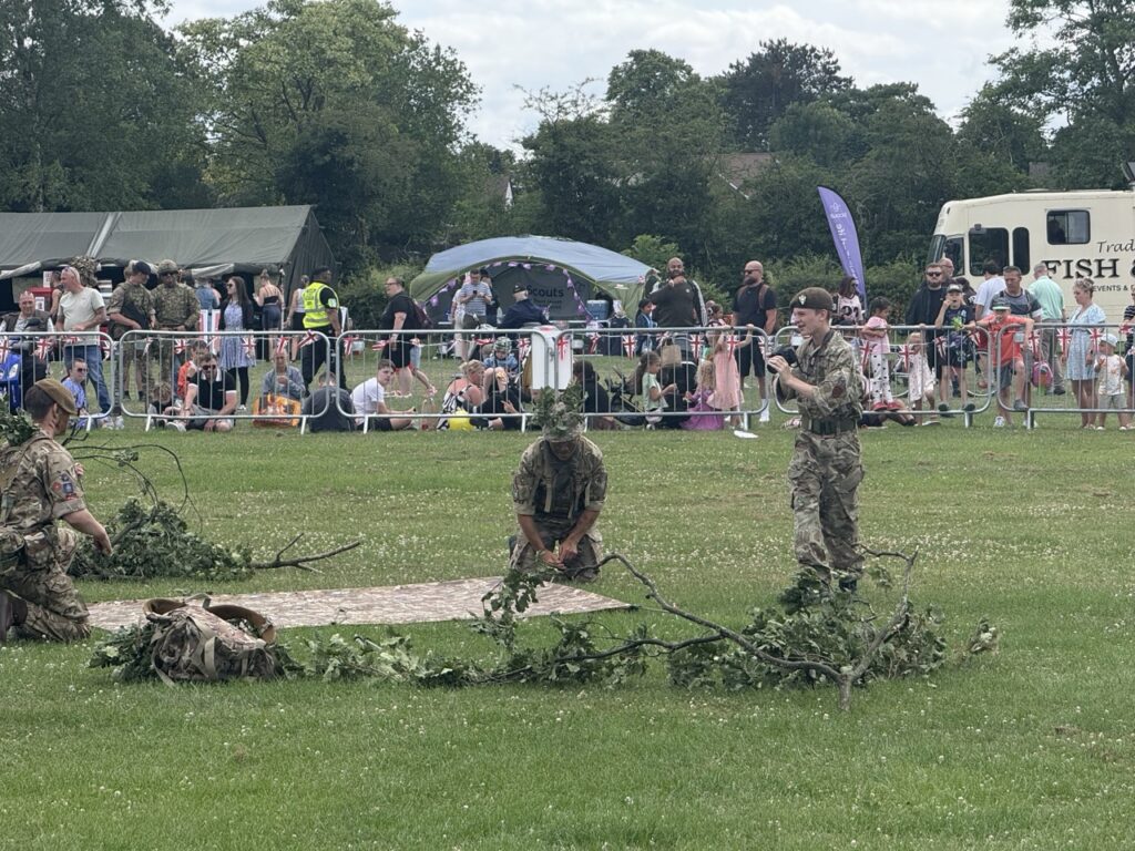 Soldiers in camouflage in a field demonstration watched by a large crowd