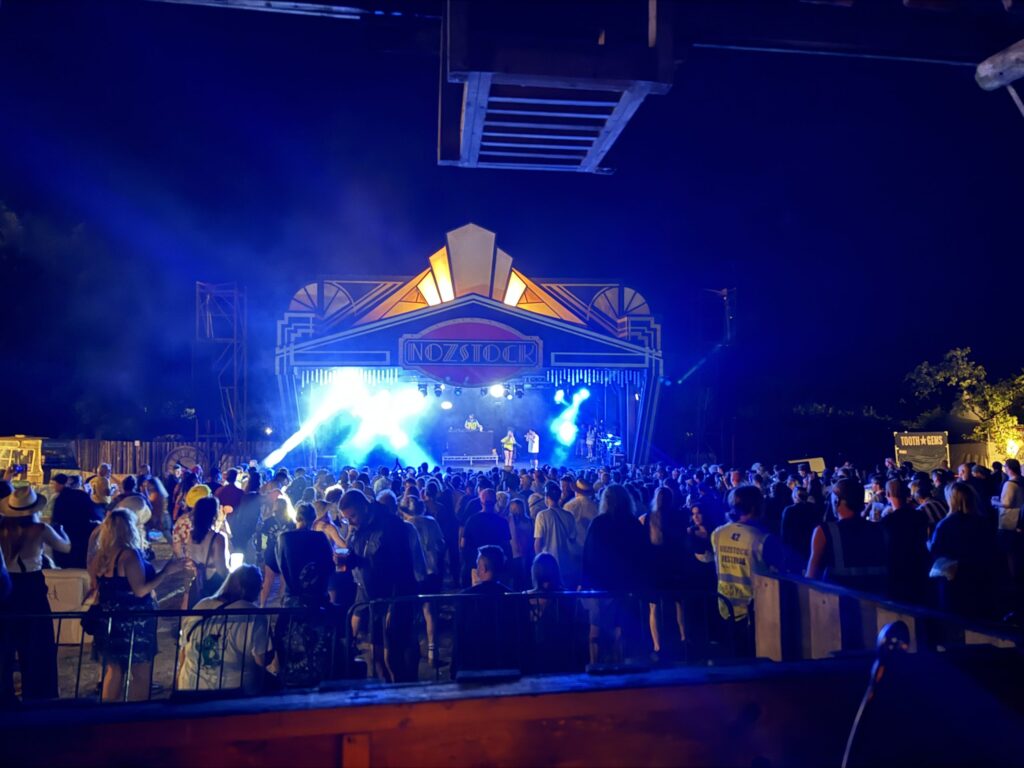 Blue-lit stage under a canopy at night with a big crowd in front
