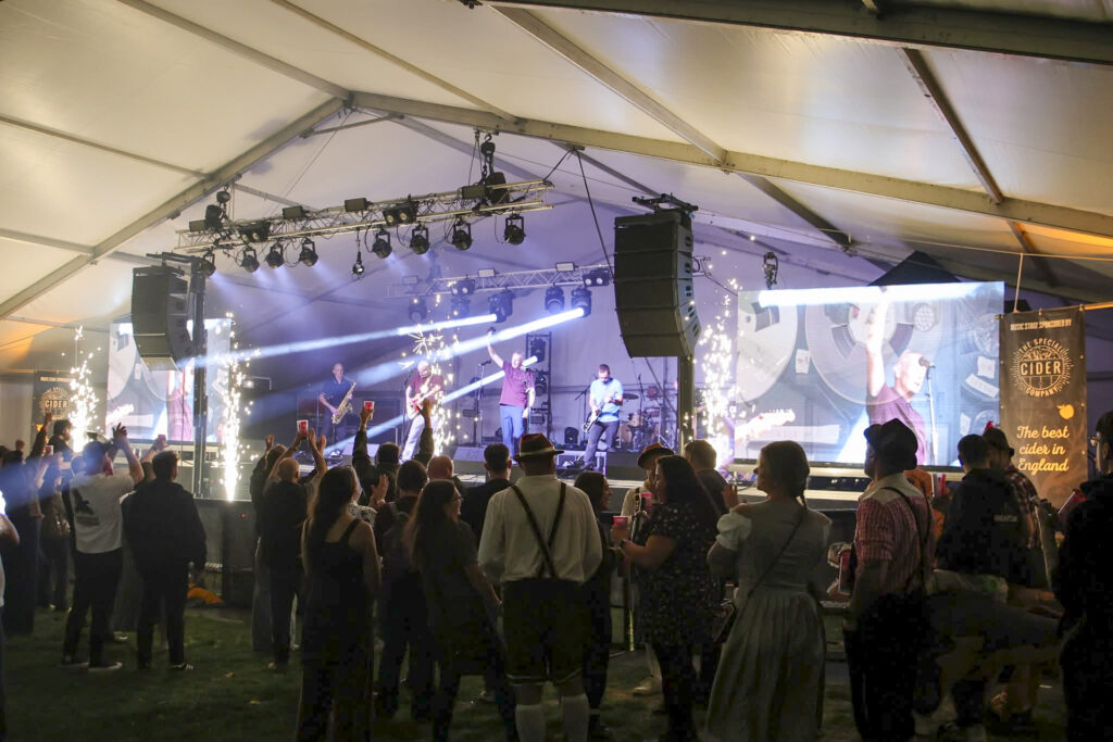 Band on stage in a large marquee with stage lighting and a crowd in front